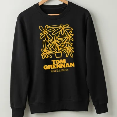 Line art illustration of a flower pot with flowers and text 'TOM GRENNAN What Is & Maybes' — printed on a unisex sweatshirt