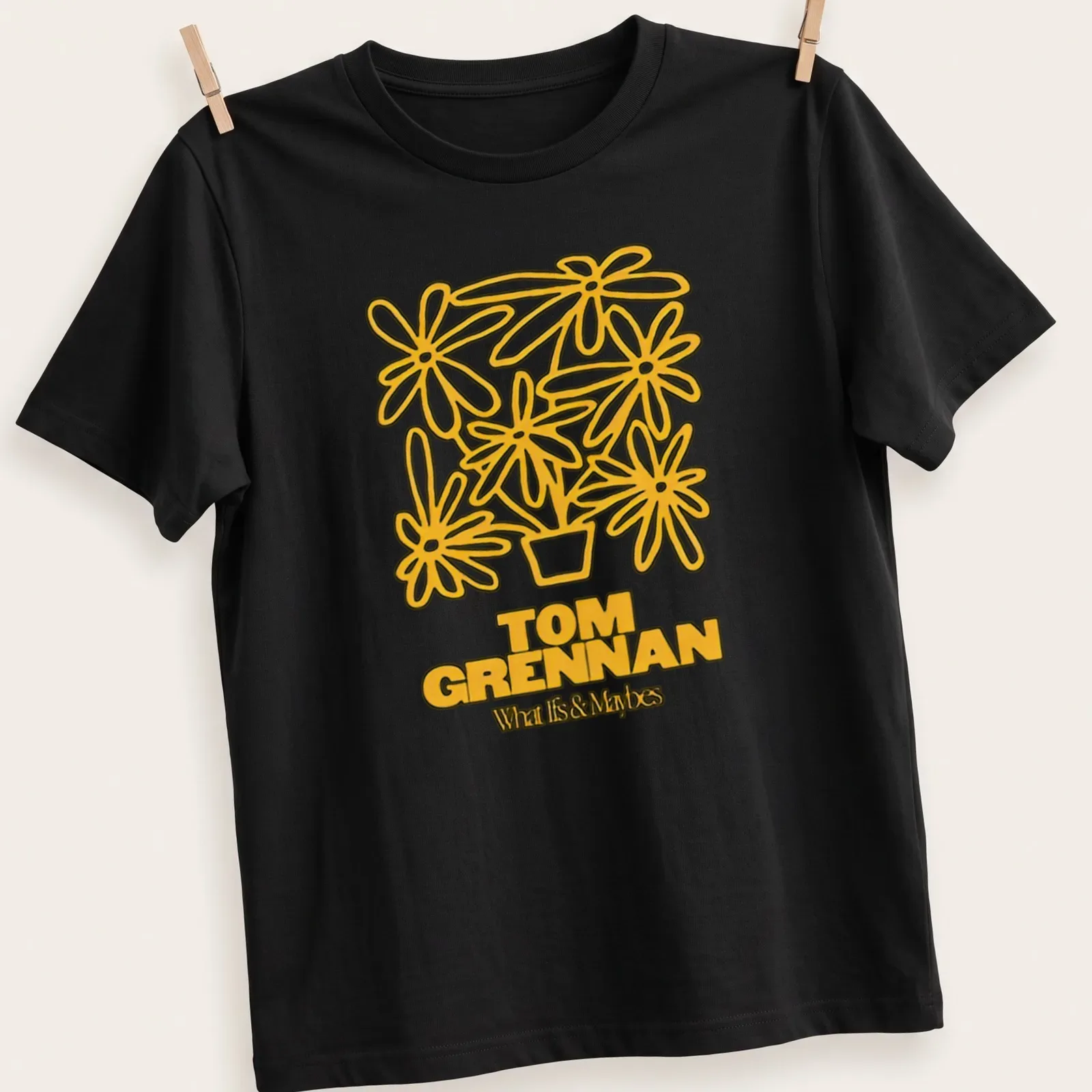 Line art illustration of a flower pot with flowers and text 'TOM GRENNAN What Is & Maybes' — printed on a unisex t-shirt